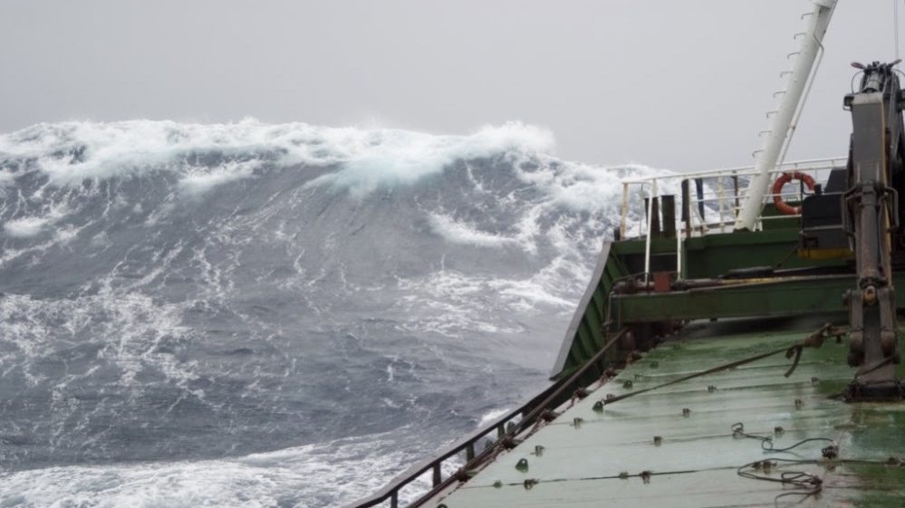 A cargo ship facing a large wave in the ocean