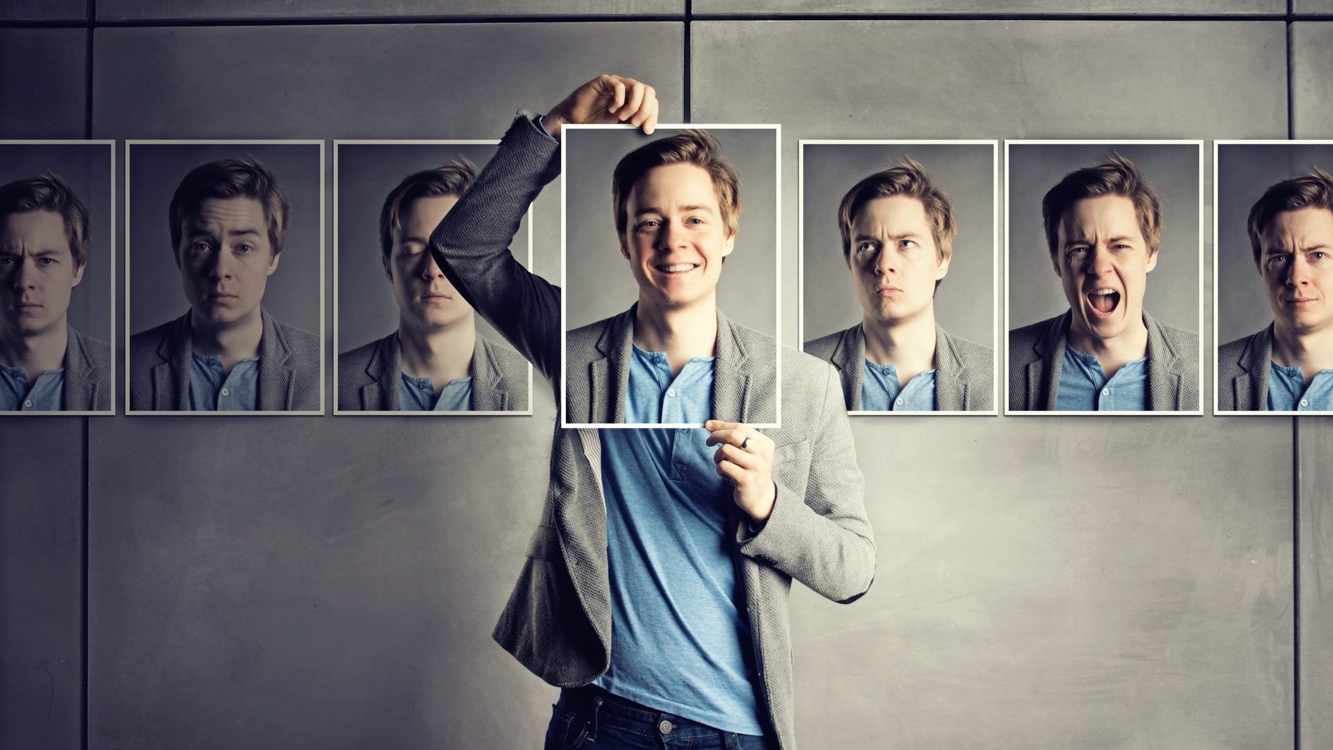 A young man holding a picture of his smiling face in front of his head with other pictures of different versions of his face lined up on a wall behind him